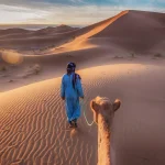Two Tuareg nomads dressed in traditional long blue robes, lead a camel through the dunes of the Sahara Desert at sunrise in Morocco.
