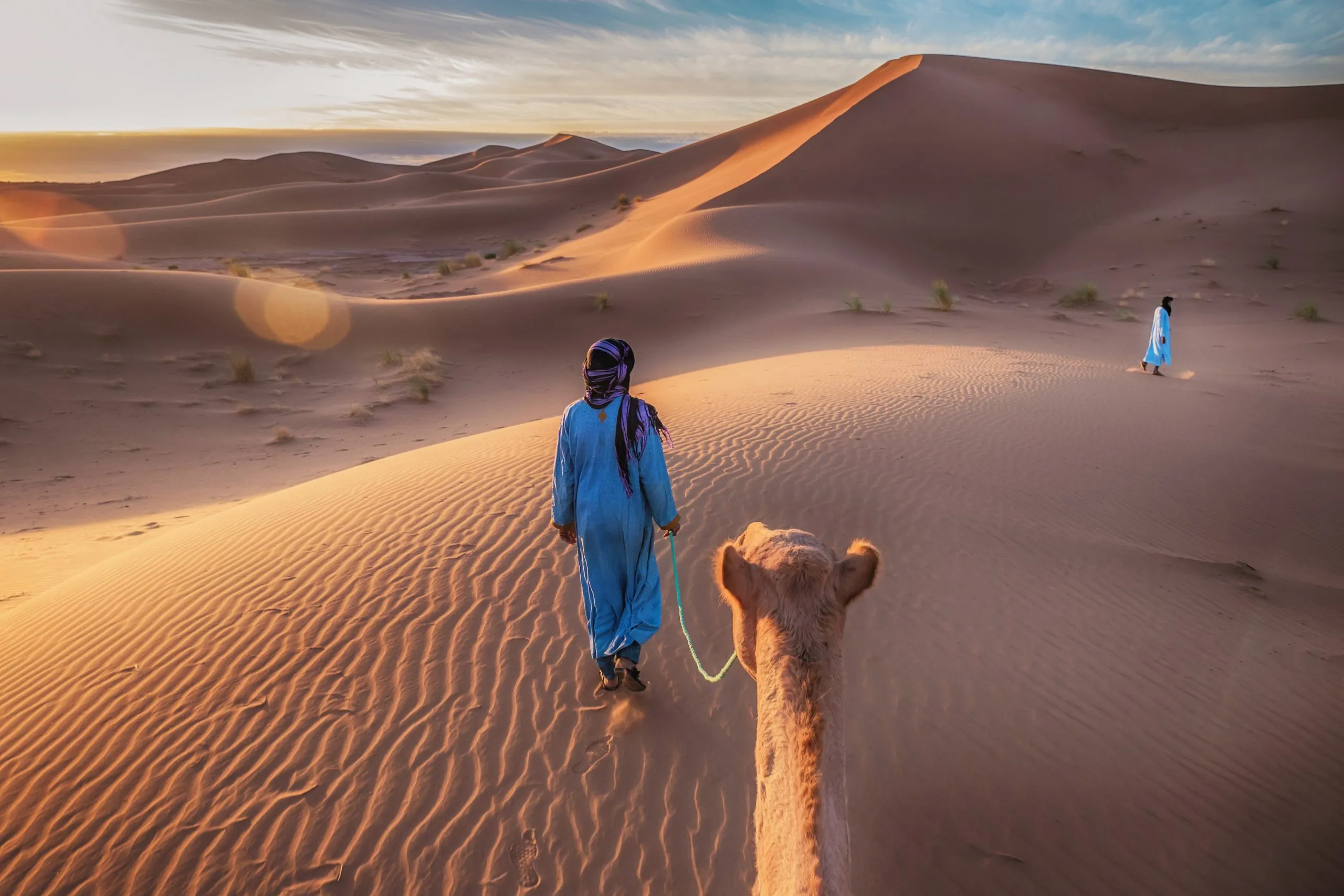 Two Tuareg nomads dressed in traditional long blue robes, lead a camel through the dunes of the Sahara Desert at sunrise in Morocco.